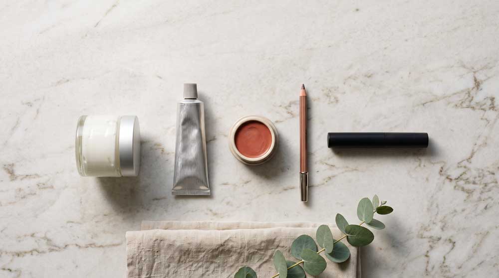 Five everyday makeup products for mature skin laid out on a counter: moisturizer, tinted sunscreen, cream blush, brow pencil, and mascara