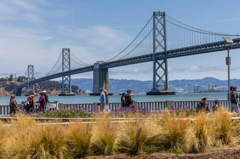 People walk by the bay bridge on a sunny day